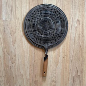 May include: A circular metal splatter screen with a dark, weathered finish and a dense pattern of small perforations. The tool features a short, light-colored wooden handle that is charred and darkened at the connection point to the metal disc.