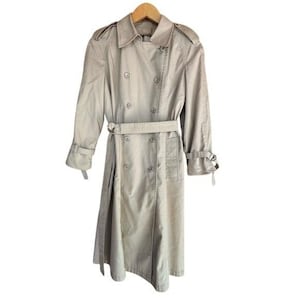 May include: A beige trench coat with a double-breasted design, a belt, and buttoned cuffs. The coat is hanging on a wooden hanger. The coat has a classic, timeless style.