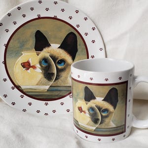 May include: A white ceramic mug and plate set featuring a Siamese cat peering through a fishbowl with a goldfish. The plate has a brown border and paw print accents. The mug and plate are a matching set.