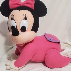 K&ouml;nnte beinhalten: Pl&uuml;sch Minnie Maus Puppe in Krabbelposition. Die Puppe hat ein rosa Outfit, eine rosa Schleife, schwarze Ohren und gro&szlig;e wei&szlig;e Augen. Wei&szlig;e Spitze an den F&uuml;&szlig;en.