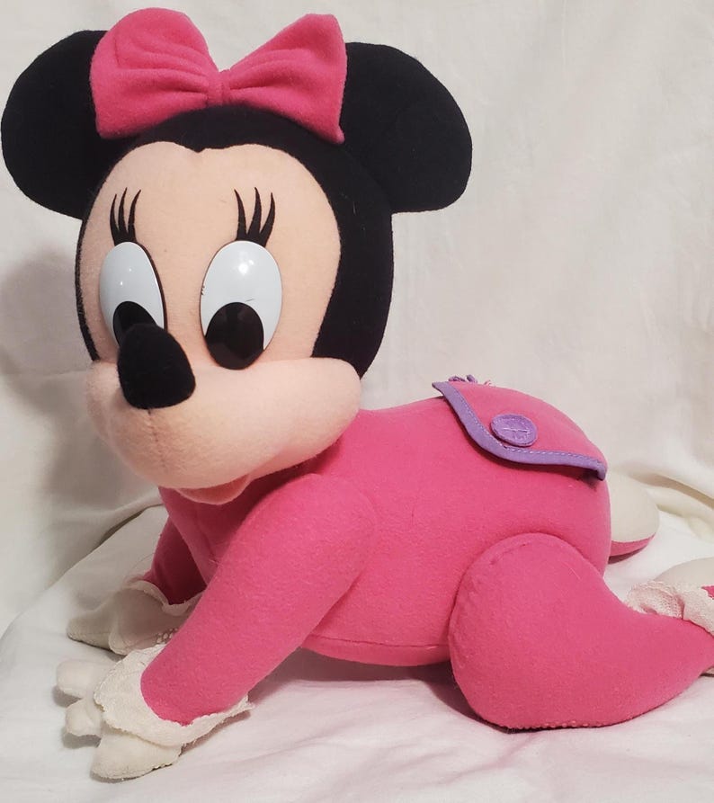 K&ouml;nnte beinhalten: Pl&uuml;sch Minnie Maus Puppe in Krabbelposition. Die Puppe hat ein rosa Outfit, eine rosa Schleife, schwarze Ohren und gro&szlig;e wei&szlig;e Augen. Wei&szlig;e Spitze an den F&uuml;&szlig;en.