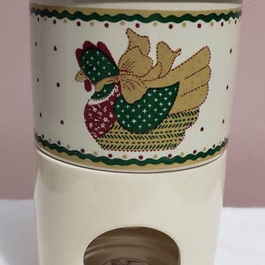 Puede incluir: Mantequillera de cerámica color crema con tapa. El plato presenta un diseño decorativo de gallina con detalles en rojo, verde y amarillo. El diseño está sobre un fondo de pequeños puntos rojos y un borde verde y amarillo.