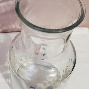 K&ouml;nnte beinhalten: Ein klarer Glaskrug mit breiter &Ouml;ffnung und rundem K&ouml;rper. Der Krug hat ein dekoratives Muster aus blauen und rosa Blumenmotiven. Geeignet zum Servieren von Getr&auml;nken.