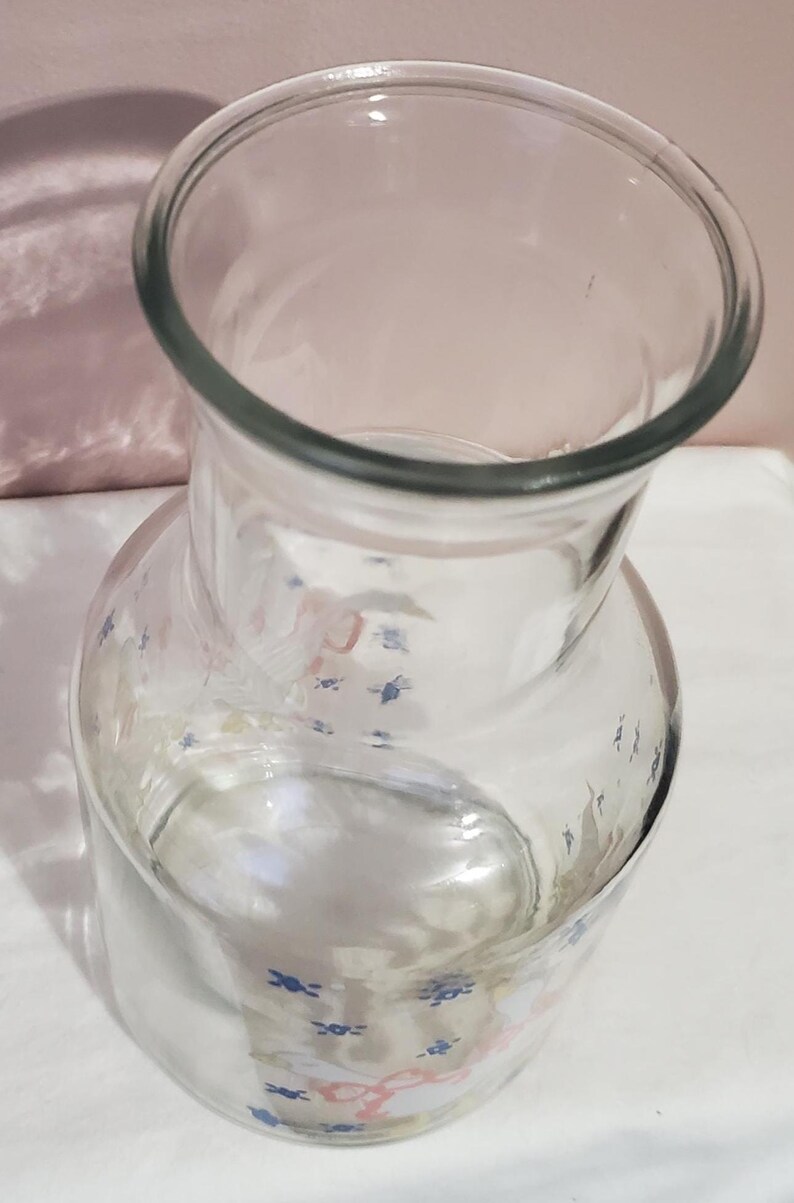 K&ouml;nnte beinhalten: Ein klarer Glaskrug mit breiter &Ouml;ffnung und rundem K&ouml;rper. Der Krug hat ein dekoratives Muster aus blauen und rosa Blumenmotiven. Geeignet zum Servieren von Getr&auml;nken.