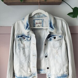 May include: Light wash denim jacket with distressed details, hanging on a white hanger. The jacket has a button-up front, two chest pockets, and the brand name "Mudd" is visible. The jacket is size XL.