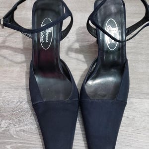 以下が含まれることがあります： ネイビーブルーのアンクルストラップ付きハイヒールサンダル。靴はスクエアトゥのデザインで、ピンヒールです。「Browns Couture」のブランド名がインソールに見えます。サンダルは、明るい灰色の木製の表面に置かれています。