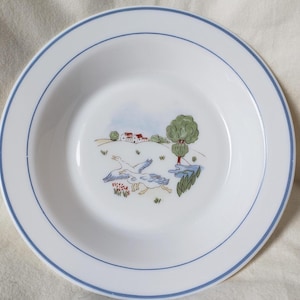 May include: White ceramic bowl with a blue rim and a decorative inner design. The design features two geese, a tree, houses, and a small pond. The bowl has a simple, elegant design.