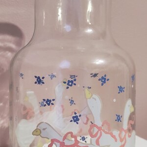 K&ouml;nnte beinhalten: Transparenter Glaskrug mit breitem Boden und schmalem Hals. Der Krug ist mit wei&szlig;en G&auml;nsen mit rosa Schleifen und blauen Blumenmotiven verziert. Die G&auml;nse haben gelbe F&uuml;&szlig;e und Schn&auml;bel. Ein dekorativer Artikel f&uuml;r die K&uuml;che.