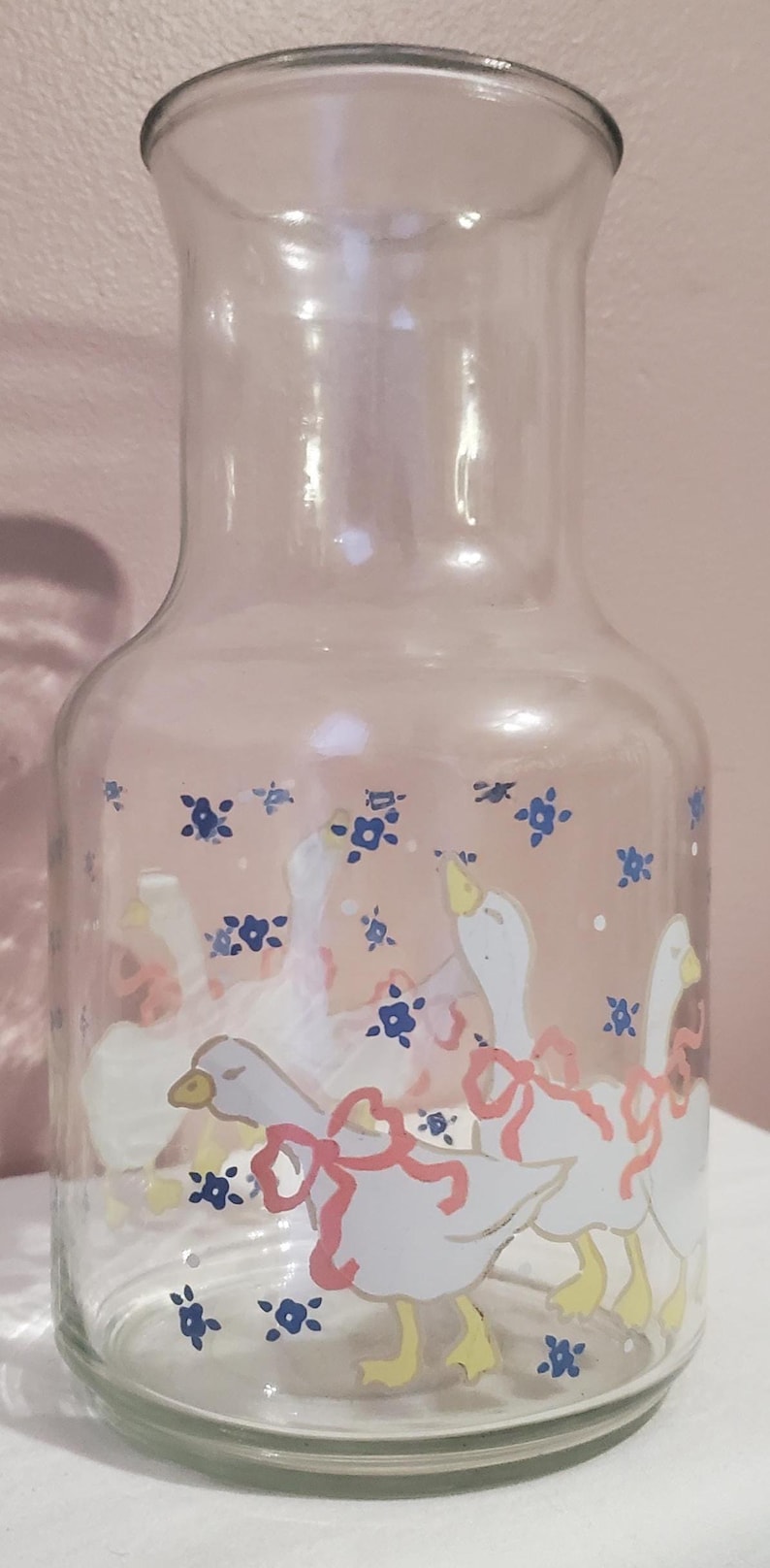 K&ouml;nnte beinhalten: Transparenter Glaskrug mit breitem Boden und schmalem Hals. Der Krug ist mit wei&szlig;en G&auml;nsen mit rosa Schleifen und blauen Blumenmotiven verziert. Die G&auml;nse haben gelbe F&uuml;&szlig;e und Schn&auml;bel. Ein dekorativer Artikel f&uuml;r die K&uuml;che.