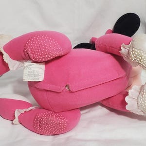 K&ouml;nnte beinhalten: Ein rosa Pl&uuml;schtier, m&ouml;glicherweise eine Figur, mit schwarzen Ohren, wei&szlig;en Handschuhen und rosa F&uuml;&szlig;en. Die F&uuml;&szlig;e haben ein strukturiertes, gepunktetes Muster. Das Spielzeug liegt auf einer wei&szlig;en Oberfl&auml;che, mit einem Etikett.