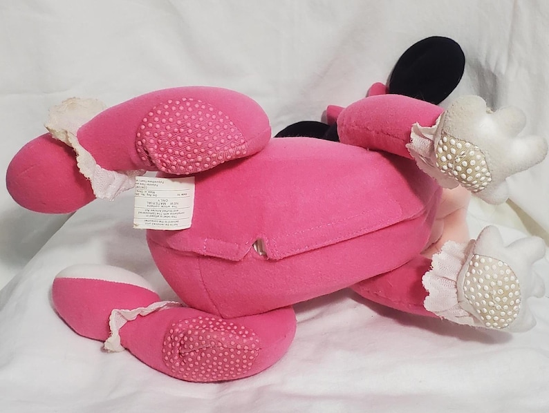 K&ouml;nnte beinhalten: Ein rosa Pl&uuml;schtier, m&ouml;glicherweise eine Figur, mit schwarzen Ohren, wei&szlig;en Handschuhen und rosa F&uuml;&szlig;en. Die F&uuml;&szlig;e haben ein strukturiertes, gepunktetes Muster. Das Spielzeug liegt auf einer wei&szlig;en Oberfl&auml;che, mit einem Etikett.