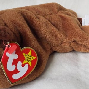 Peut inclure: Une peluche de castor marron avec une longue queue plate et de petites oreilles. Le jouet a une étiquette rouge en forme de cœur avec du texte blanc et une étoile jaune. Le castor a des yeux noirs et des dents blanches.