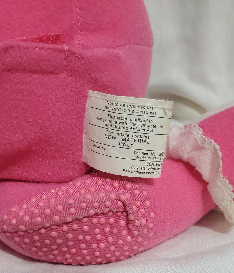 K&ouml;nnte beinhalten: Nahaufnahme eines rosa Pl&uuml;schtiers mit einem Etikett, das "NEUES MATERIAL NUR" angibt. Der Fu&szlig; des Spielzeugs hat ein strukturiertes, gepunktetes Muster. Hergestellt in China.