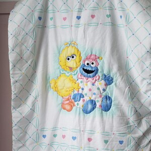 May include: White baby blanket featuring Big Bird and Cookie Monster appliqu&eacute;s. The blanket has a repeating pattern of hearts and a diamond grid design in pastel colors. The characters are in the center of the blanket.