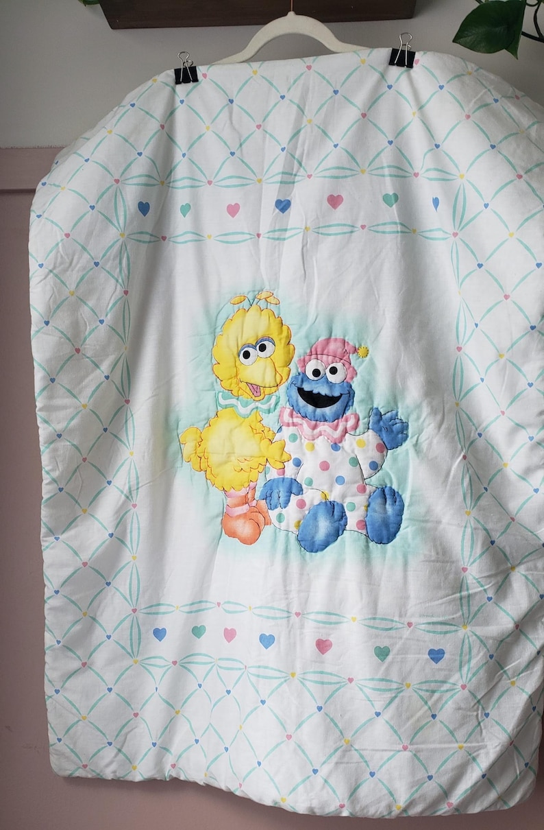 May include: White baby blanket featuring Big Bird and Cookie Monster appliqu&eacute;s. The blanket has a repeating pattern of hearts and a diamond grid design in pastel colors. The characters are in the center of the blanket.