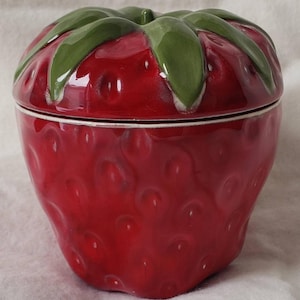 May include: A ceramic strawberry-shaped container with a lid. The container is a vibrant red with a textured surface, mimicking the appearance of a real strawberry. The lid features green leaves, adding to the realistic design. The container is likely used for storage.