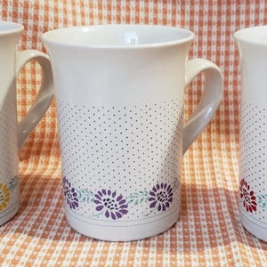 Puede incluir: Tres tazas de cerámica blanca con un estampado de lunares y diseños florales. Cada taza tiene un borde floral diferente: amarillo, morado y rojo. Las tazas tienen un asa curva y una parte superior ligeramente acampanada.
