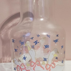 K&ouml;nnte beinhalten: Transparenter Glaskrug mit einem dekorativen Design aus wei&szlig;en G&auml;nsen mit gelben F&uuml;&szlig;en und rosa Schleifen. Blaue Blumenmotive sind um die G&auml;nse herum verstreut. Der Krug hat eine klassische Form und eignet sich zum Servieren von Getr&auml;nken.