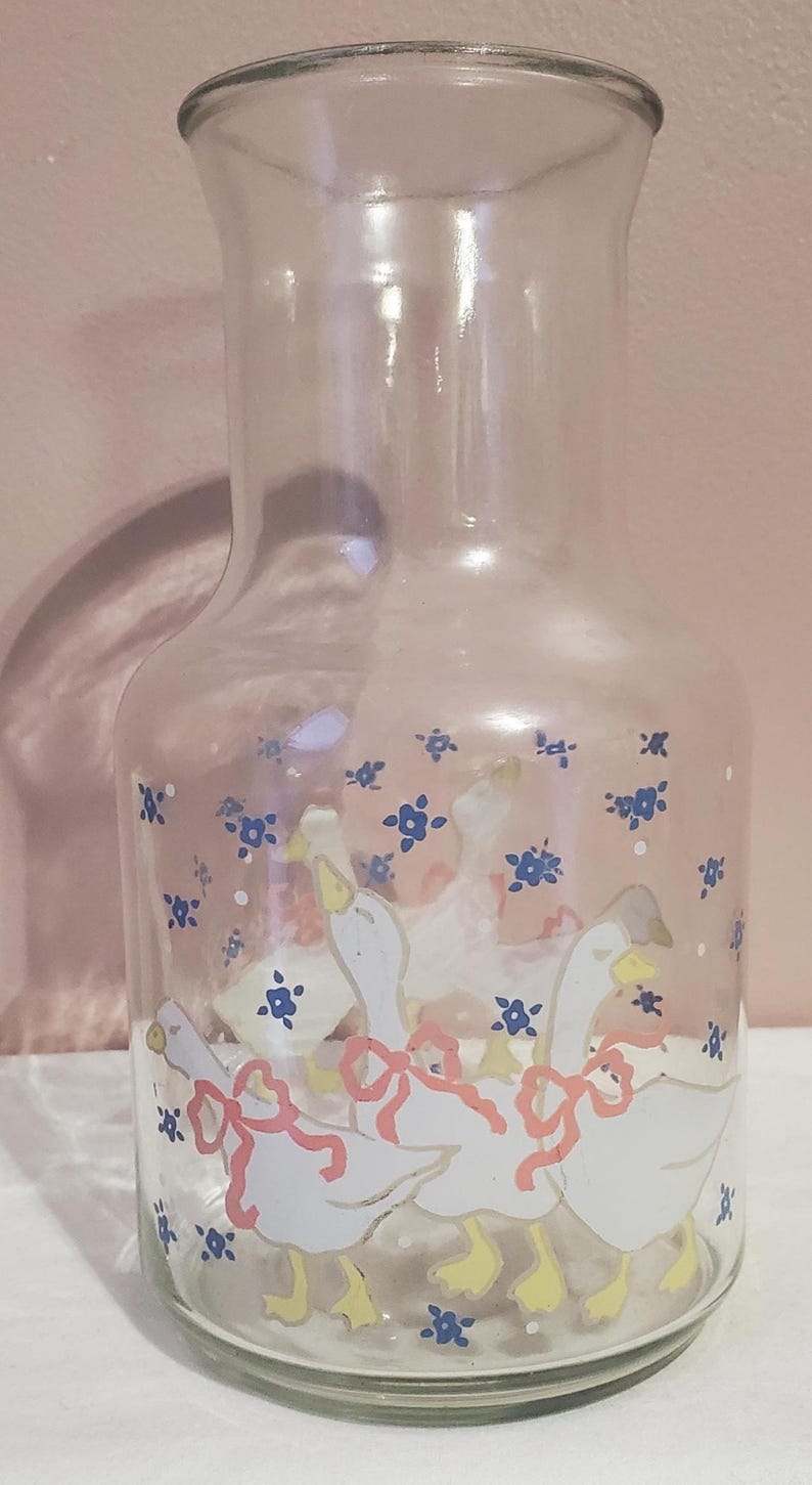 K&ouml;nnte beinhalten: Transparenter Glaskrug mit einem dekorativen Design aus wei&szlig;en G&auml;nsen mit gelben F&uuml;&szlig;en und rosa Schleifen. Blaue Blumenmotive sind um die G&auml;nse herum verstreut. Der Krug hat eine klassische Form und eignet sich zum Servieren von Getr&auml;nken.