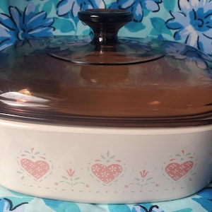 May include: A white rectangular casserole dish with two side handles and a dark brown translucent glass lid. The exterior of the dish features a repeating pattern of pink hearts and small green floral accents along the base of the vessel.