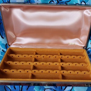May include: A rectangular jewelry box with a hinged lid, featuring a smooth, light pink satin interior lining and a mustard yellow velvet base with three rows of scalloped slots for rings. The brand name MELE is printed in gold on the satin.