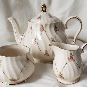Könnte beinhalten: Ein cremefarbenes Vintage-Teeservice mit Teekanne, Sahnekännchen und Zuckerdose. Das Set hat ein Spiraldesign, rosa Rosenakzente und goldfarbene Verzierungen. Die Teekanne hat einen goldenen Knauf und Griff.