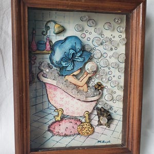 Puede incluir: Una obra de arte enmarcada que representa a una persona en una bañera, con un sombrero azul, rodeada de burbujas. La bañera rosa tiene un estampado floral. El marco es de madera. La obra incluye un cabezal de ducha y objetos decorativos.