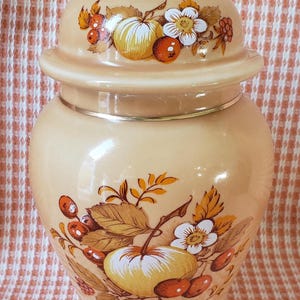 May include: A vintage beige ceramic jar with a lid, adorned with a fruit and floral design. The jar features a gold-colored finial on the lid and a silver band around the middle. The fruit and floral design includes apples, berries, and white flowers.