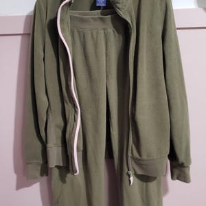 Peut inclure: Ensemble de survêtement en polaire vert olive, comprenant une veste zippée avec une fermeture éclair rose et un pantalon assorti. L'ensemble est suspendu à un cintre blanc contre un mur rose clair. La veste a un col montant et le pantalon une taille élastique.