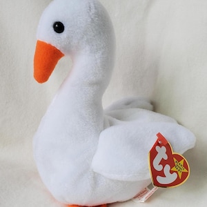 Peut inclure: Une peluche blanche en forme d'oie avec un bec et des pieds orange. L'oie a un œil noir et une étiquette rouge attachée à son aile. L'étiquette porte les mots "BEANIE BABY" et une forme de cœur.