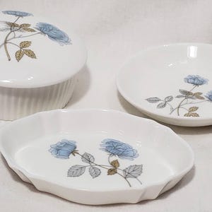 Peut inclure: Un ensemble de trois plats en céramique blanche. L'ensemble comprend un récipient rond avec couvercle, un petit bol rond et un plat ovale. Chaque pièce est décorée d'un motif de rose bleue avec des feuilles grises et des tiges brunes.