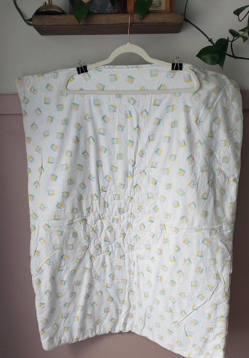 May include: White baby blanket with a repeating pattern of colorful letter blocks. The blocks feature the letters A, B, and C in pastel colors. The blanket is hanging on a white hanger.