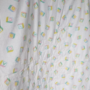 May include: White baby blanket with a repeating pattern of colorful alphabet blocks. Each block features a letter (A, B, or C) in a pastel color scheme of pink, yellow, and green. The blanket is hanging.