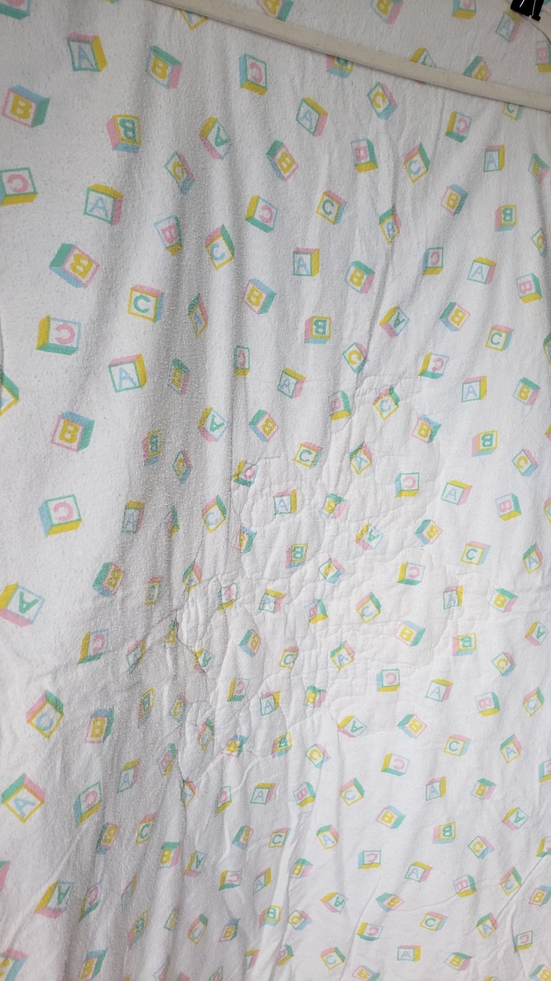 May include: White baby blanket with a repeating pattern of colorful alphabet blocks. Each block features a letter (A, B, or C) in a pastel color scheme of pink, yellow, and green. The blanket is hanging.