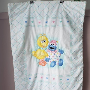 May include: White baby quilt featuring Big Bird, Cookie Monster, and a smaller Big Bird. The quilt has a diamond pattern with hearts in pink, blue, and green. The characters are in the center of the quilt.