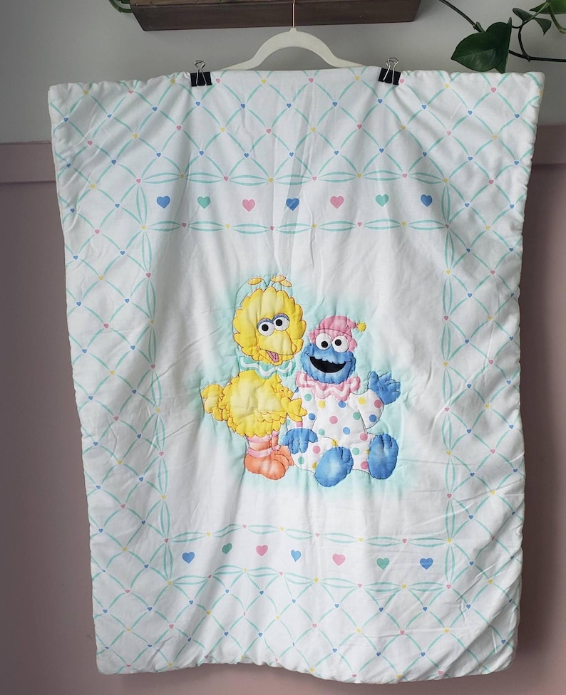 May include: White baby quilt featuring Big Bird, Cookie Monster, and a smaller Big Bird. The quilt has a diamond pattern with hearts in pink, blue, and green. The characters are in the center of the quilt.