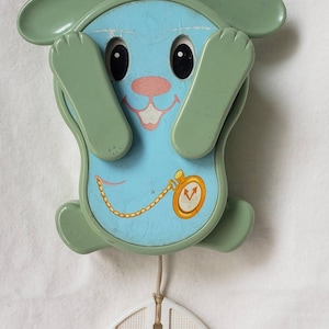 May include: A vintage baby mobile featuring a cartoon bunny design. The bunny has a light blue face with black eyes, a pink nose, and a smiling mouth. The bunny's ears and arms are green. A gold-colored pocket watch is on the bunny's chest.