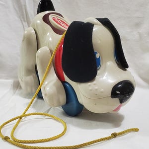 May include: A vintage Playskool pull-along dog toy. The toy dog is mainly off-white with black ears, a black nose, and blue eyes. It has a red and blue accent on its side and a yellow pull string. The toy is made of plastic.