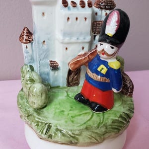 May include: A ceramic figurine featuring a castle and a soldier. The castle is white with brown accents, and the soldier wears a blue jacket, red pants, and a black hat. The base is white with green grass.