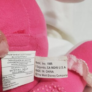 K&ouml;nnte beinhalten: Nahaufnahme eines rosa Pl&uuml;schtiers mit wei&szlig;er Verzierung. Das Spielzeug hat einen strukturierten Boden und ein Etikett mit der Aufschrift "Mattel, Inc 1995, Made in China, The Walt Disney Company."