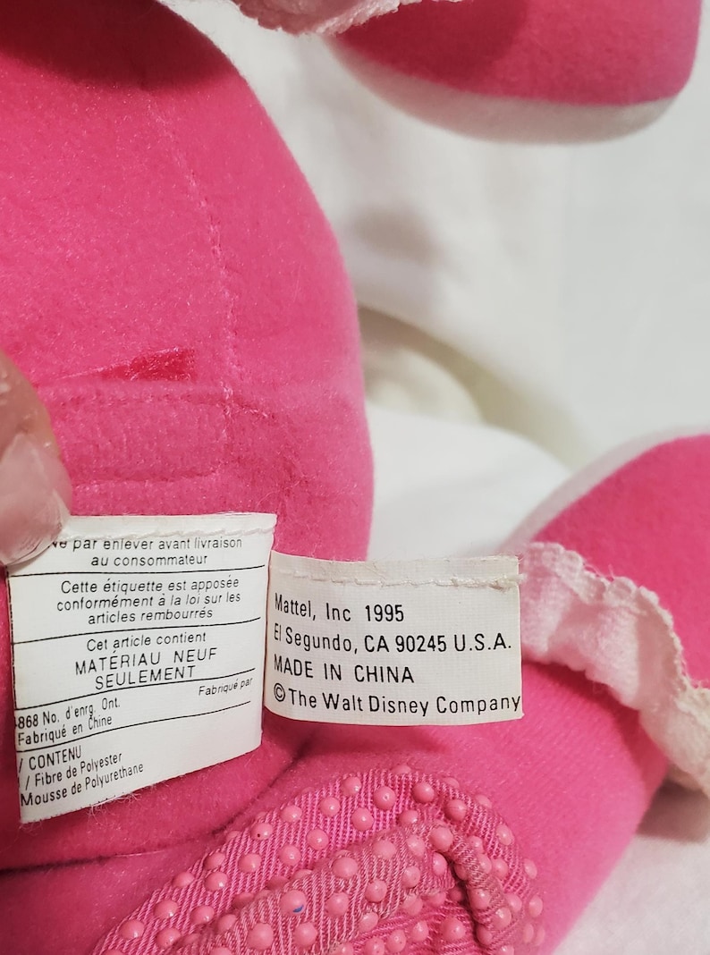 K&ouml;nnte beinhalten: Nahaufnahme eines rosa Pl&uuml;schtiers mit wei&szlig;er Verzierung. Das Spielzeug hat einen strukturierten Boden und ein Etikett mit der Aufschrift "Mattel, Inc 1995, Made in China, The Walt Disney Company."