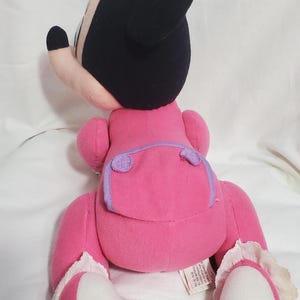 K&ouml;nnte beinhalten: Ein Pl&uuml;sch-Minnie-Mouse-Puppe mit einem rosa Kleid, einer Schleife und Schuhen sowie schwarzen Ohren und einem Kopf. Die Puppe hat eine lila Windel und wei&szlig;e Spitzentrimm an den Schuhen. Die Puppe ist aus weichem Stoff.