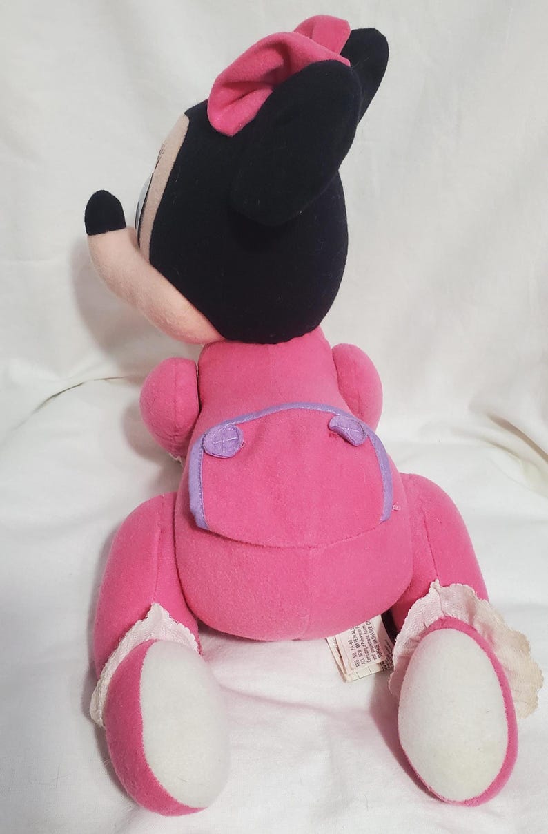 K&ouml;nnte beinhalten: Ein Pl&uuml;sch-Minnie-Mouse-Puppe mit einem rosa Kleid, einer Schleife und Schuhen sowie schwarzen Ohren und einem Kopf. Die Puppe hat eine lila Windel und wei&szlig;e Spitzentrimm an den Schuhen. Die Puppe ist aus weichem Stoff.