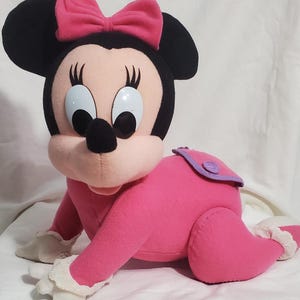 K&ouml;nnte beinhalten: Ein Pl&uuml;sch Minnie Maus Puppe in Krabbelstellung. Die Puppe hat ein rosa Outfit, eine rosa Schleife und schwarze Ohren. Das Gesicht der Puppe hat gro&szlig;e, blaue Augen und eine schwarze Nase. Die Puppe hat einen lila Windel&uuml;berzug.
