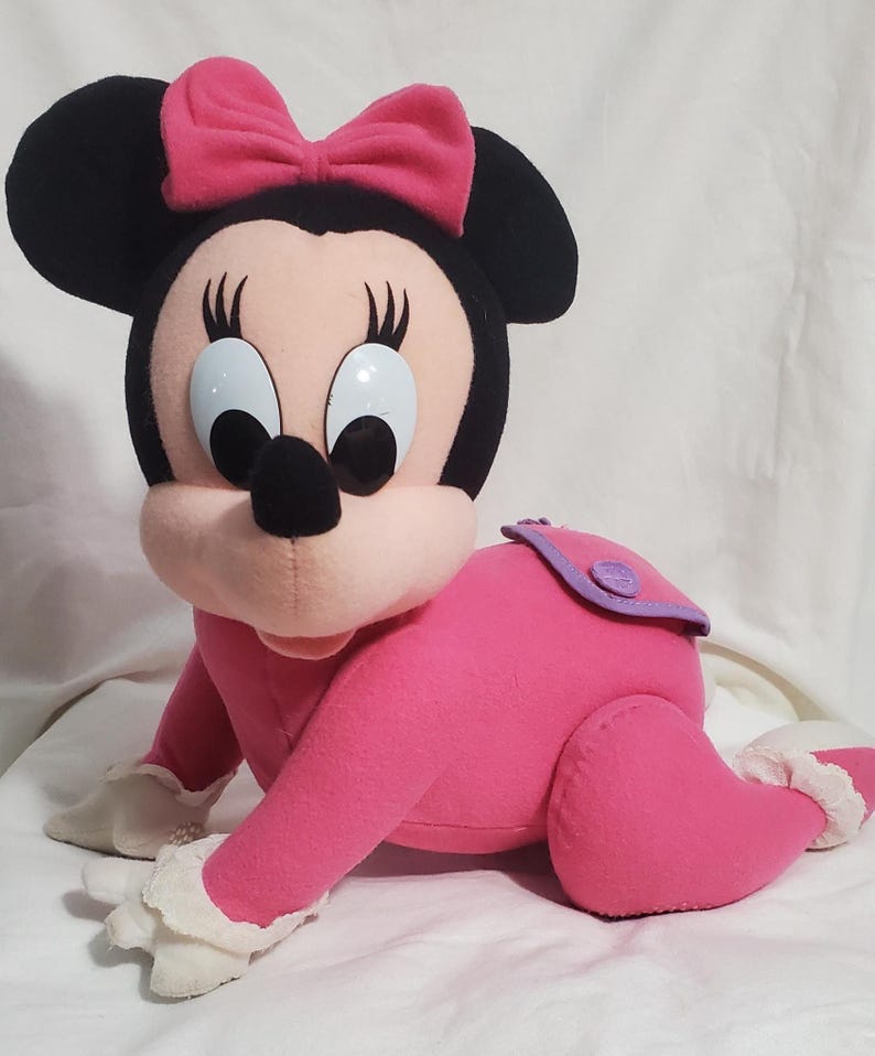 K&ouml;nnte beinhalten: Ein Pl&uuml;sch Minnie Maus Puppe in Krabbelstellung. Die Puppe hat ein rosa Outfit, eine rosa Schleife und schwarze Ohren. Das Gesicht der Puppe hat gro&szlig;e, blaue Augen und eine schwarze Nase. Die Puppe hat einen lila Windel&uuml;berzug.
