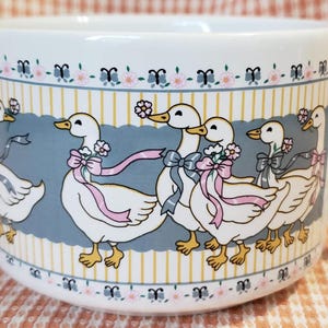May include: White ceramic mug with two goose-shaped handles. The mug features a repeating pattern of geese with pink ribbons and flowers, set against a blue and yellow striped background. The rim has a floral and butterfly design.