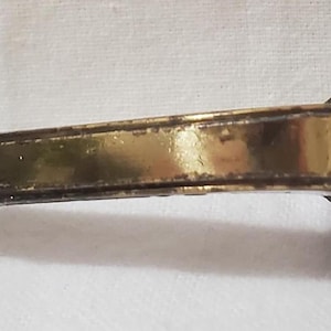 May include: A decorative bronze-toned cabinet pull with a gold-colored handle. The pull features ornate detailing on the ends and a rectangular gold-colored bar in the center. The hardware is designed for furniture.