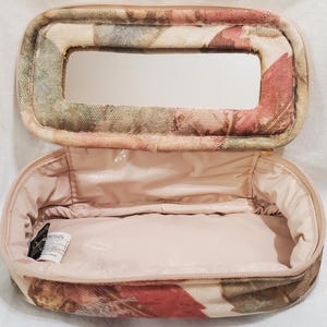 May include: Open cosmetic bag with a floral pattern and a mirror in the lid. The bag has a rectangular shape with rounded edges and a zip closure. The interior is pale pink, and the exterior features a pattern of red, green, and beige leaves.
