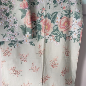 Pode incluir: Tecido floral com fundo bege, apresentando grandes rosas rosa e pêssego, flores brancas e folhas verdes. A parte inferior tem um padrão de pequenas flores cor de pêssego. O tecido está pendurado em um cabide branco.