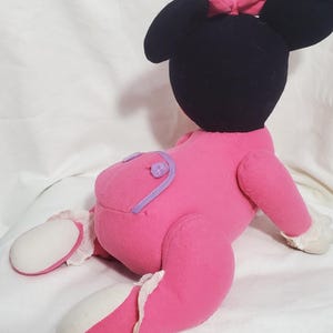 K&ouml;nnte beinhalten: Eine Pl&uuml;schpuppe von Minnie Mouse, die auf der Seite liegt. Die Puppe hat einen schwarzen Kopf mit gro&szlig;en Ohren und einer rosa Schleife. Sie tr&auml;gt ein rosa Outfit mit lila Akzenten und wei&szlig;er Verzierung an den F&uuml;&szlig;en.